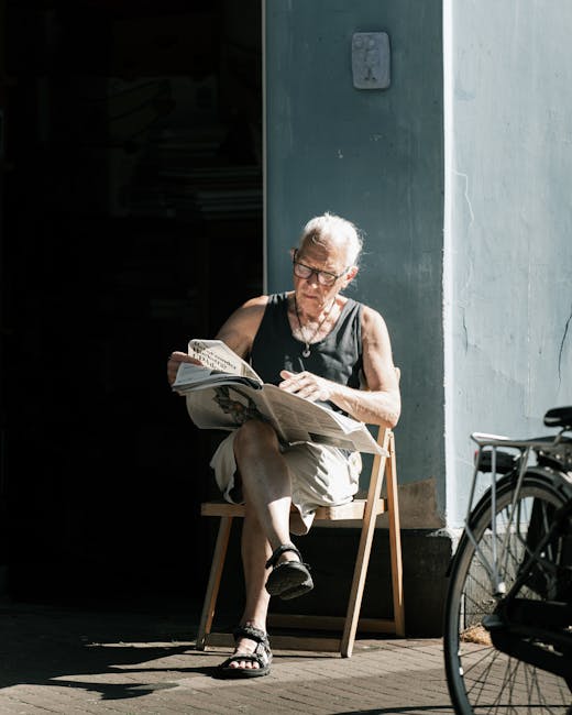 An elderly woman with short white hair and glasses is sitting on a small wooden chair outside a building with a smooth, light gray wall. She is wearing a black sleeveless top, beige shorts, and black 